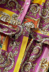 Vêtement boutonné avec des rayures éclatantes jaune, rose et orange, décoré de motifs floraux baroques et d'arabesques dorées et blanches ornés.