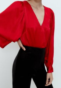Blusa in raso rosso con maniche lunghe e a sbuffo, scollatura a V profonda e dettagli arricciati alle spalle, abbinata a pantaloni neri a vita alta.