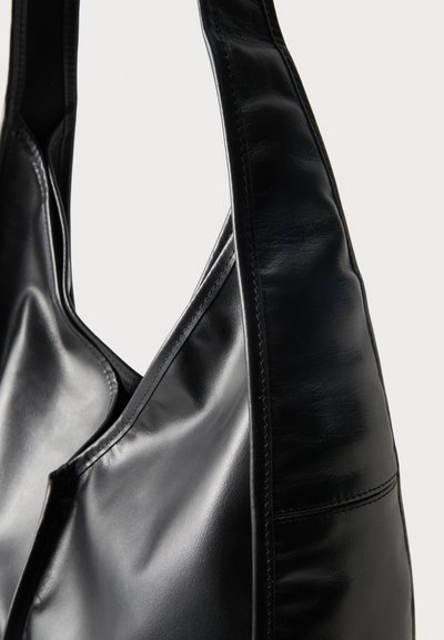 Λεπτή μαύρη δερμάτινη τσάντα ώμου με φαρδιά λουριά και εμφανείς ραφές, κοντινή λήψη σε απλό ανοιχτό φόντο.