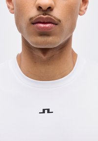 Κοντινό πλάνο του κάτω μέρους του προσώπου και του λαιμού ενός άνδρα που φοράει λευκό πουκάμισο με ένα μικρό μαύρο γεωμετρικό λογότυπο στο στήθος.