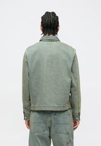 Φθαρμένο πράσινο τζιν μπουφάν με κανονικό γιακά, μανσέτες με κουμπιά και πίσω τσέπες, με χαλαρή γραμμή και ορατές λεπτομέρειες ραφής.