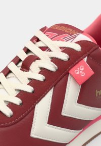 Red and white sneaker with a textured upper, featuring a prominent chevron design, white laces, and a pink accent tag on the side.