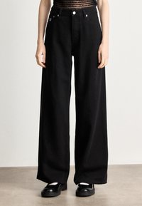 Jeans pretos de perna larga feitos de denim, apresentando bolsos frontais, uma cintura alta e um acabamento limpo. Combinados com sapatos pretos tipo loafers.