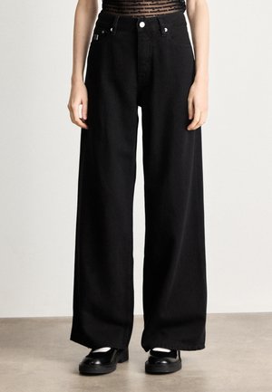 Jean noir large en denim, doté de poches avant, d'une taille haute et d'une finition soignée. Associé à des mocassins noirs.