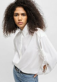 White oversized shirt with a pointed collar, long sleeves featuring tie details, and a subtle embroidered logo on the chest.