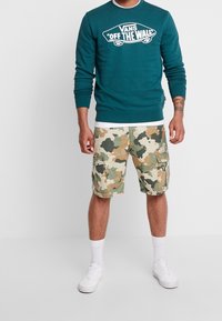 Sudadera color teal con el logo blanco "VANS OFF THE WALL", combinada con pantalones cortos de camuflaje en verde, marrón y beige, y llevada con zapatillas blancas.