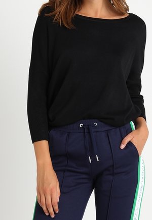 Schwarzer Strickpullover mit Bootsausschnitt und gerippten Ärmeln, kombiniert mit marineblauen Jogginghosen mit grünen Seitenstreifen und einer Kordelzugtaillierung.