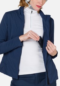 Navyfarbene Radjacke mit tailliertem Schnitt, aus leichtem Material gefertigt. Verfügt über Reißverschluss-Taschen und einen hohen Kragen, die über einem weißen Oberteil getragen wird.