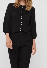 Blusa nera in maglia con maniche lunghe, caratterizzata da una chiusura frontale con bottoni e bottoni bianchi. Il tessuto ha una leggera lucentezza e un fit rilassato.