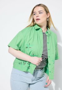Camicia verde a maniche corte con bottoni e ricami floreali, abbinata a una maglietta a righe bianco e nero e jeans azzurri chiari con dettagli di perle.