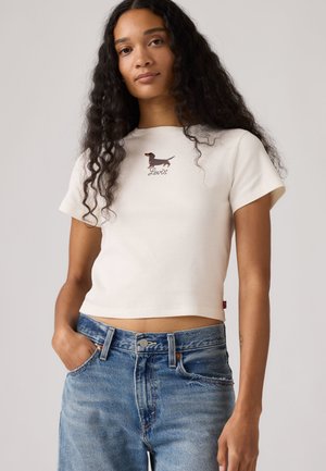 Kvinde med langt krøllet hår iført en hvid kort T-shirt med en lille gravhund og "Levi's"-logo, kombineret med blå jeans.
