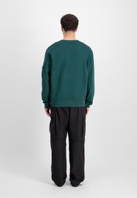 Zeleni pamučni dukserica s dugim rukavima, okruglim izrezom i rebrastim manžetama, u kombinaciji s crnim cargo hlačama koje imaju bočne džepove.