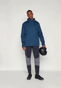 Jūras zila ūdensnecaurlaidīga jakas ar kapuci, kopā ar tumšiem biksēm ar pelēkiem akcentiem. Melna ķivere un melni apavi.