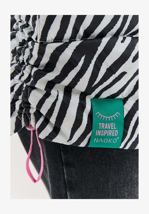 Černobílá zebrová potisková tkanina s nařaseným švem a zelenou etiketou s nápisem "Travel Inspired Naoko" připevněnou k tmavému denimu.