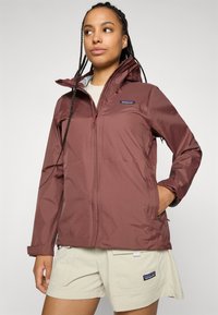 Vrouw met gevlochten haar, die een bordeauxrode Patagonia-jas en beige Patagonia-shorts draagt, en staat met één hand in de zak van de jas.