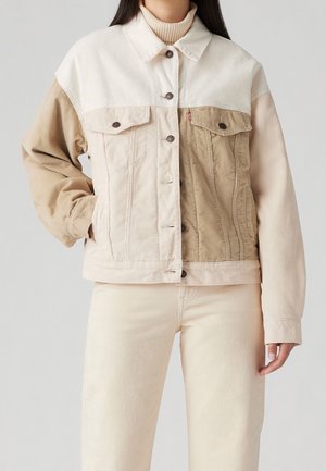 Femme portant une veste en velours côtelé bicolore beige et crème boutonnée, à manches longues, et un pantalon beige clair sur un fond uni.
