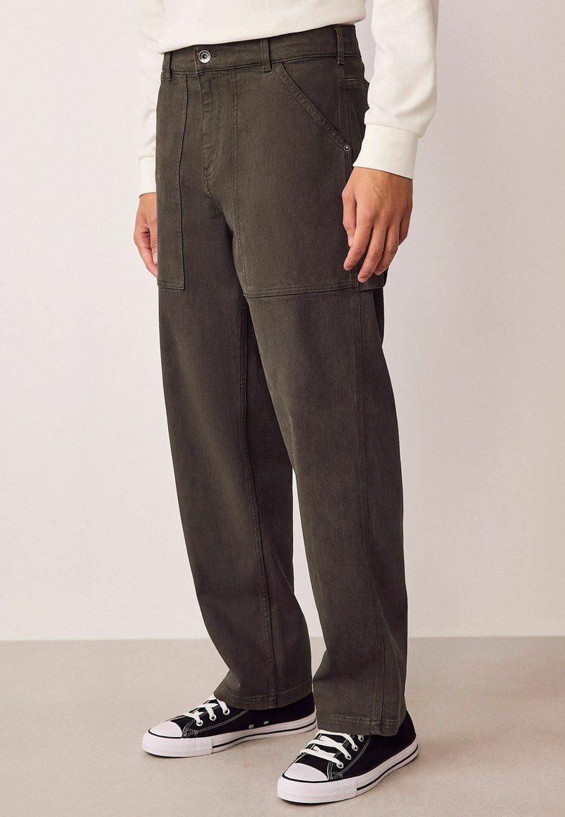 Pantalons larges en denim vert foncé, dotés de coutures doubles, de poches latérales et d'une fermeture à boutons, associés à des baskets noires.