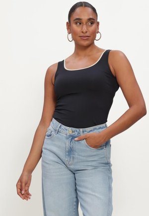 Woman with hoop earrings wearing black sleeveless top and light blue high-waisted jeans, hand in pocket, looking slightly to the side.