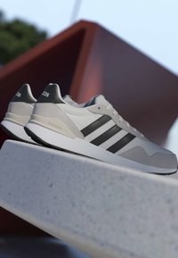 Baskets Adidas grises et blanches avec des rayures noires reposant sur une surface en béton, structure géométrique rouge floue en arrière-plan.