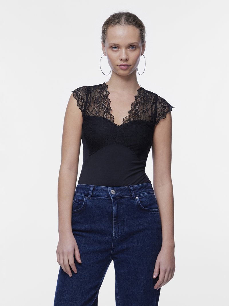 Jeune femme aux cheveux bouclés portant un haut en dentelle noire et un jean bleu foncé taille haute, debout devant un fond blanc, regardant droit devant.