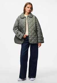 Giacca imbottita grigia con un ampio collo, bottoni a pressione e tasche laterali; indossata sopra un top beige e abbinata a jeans blu scuro e sneaker.