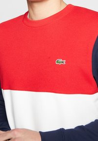 Czerwony, teksturowany sweter z białym, solidnym pasem poziomym i granatowymi rękawami. Posiada małe zielone logo krokodyla na piersi.