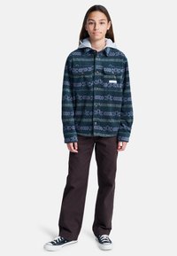 Mönstrad flanellskjorta med knäppning i mörk teal med grå huva; har abstrakta mönster och lång ärm, tillsammans med mörka byxor och sneakers.