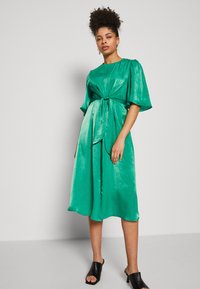 Grünes Satin-Kleid mit kurzen, puffärmeligen Ärmeln und einer gebundenen Taillendetail. Der Rock reicht bis zur Mitte der Wade und wird mit schwarzen offenen Mules kombiniert.