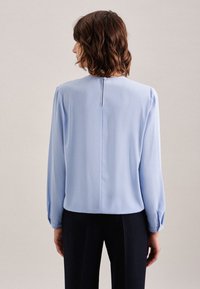Blusa a maniche lunghe azzurro chiaro con scollatura rotonda, chiusura con bottoni sul retro e tessuto morbido. Orlo arrotondato e maniche leggermente voluminoso.