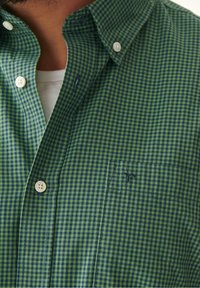 Camisa de cuadros verde y azul con cuello abotonado, de material de algodón, que presenta un bolsillo en la parte frontal y un logo bordado. Diseño de corte clásico.