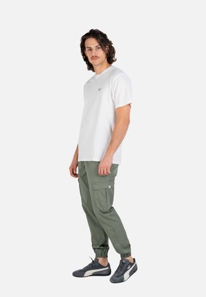 Joven con cabello ondulado que lleva una camiseta blanca, pantalones cargo color verde oliva y zapatillas negras Puma, posando contra un fondo blanco liso.