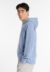 Hoodie azzurro chiaro realizzato in tessuto morbido, con tasca frontale e cappuccio con cordoncino. Modello mostrato di profilo su uno sfondo bianco.