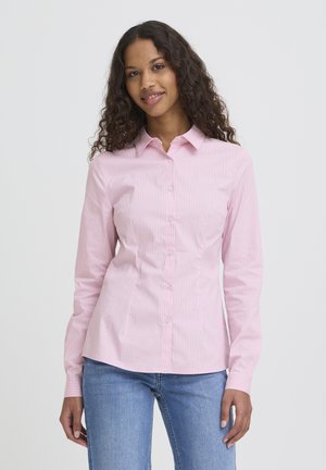 Kvinde med krøllet hår iført en tætsiddende lyserød skjorte med knapper og blå jeans, der står foran en ensfarvet lys baggrund.