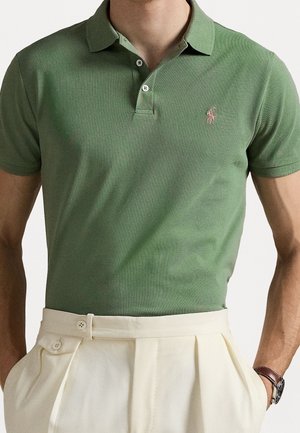 Mand iført en grøn kortærmet polo tucked ind i højtaljede cremefarvede plisserede bukser med ur på venstre håndled.