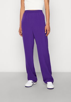 Person wearing purple loose pants, white sneakers with purple stripes, and a white cropped top, standing against a plain light background.
