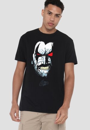 Junger Mann trägt ein schwarzes T-Shirt mit einer Grafik eines comicartigen Charakters mit weißem Gesicht, roten Augen und grimmigem Ausdruck auf der Vorderseite.