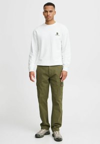 White long-sleeve shirt with small green logo, paired with olive green cargo pants featuring side pockets and grey sneakers.