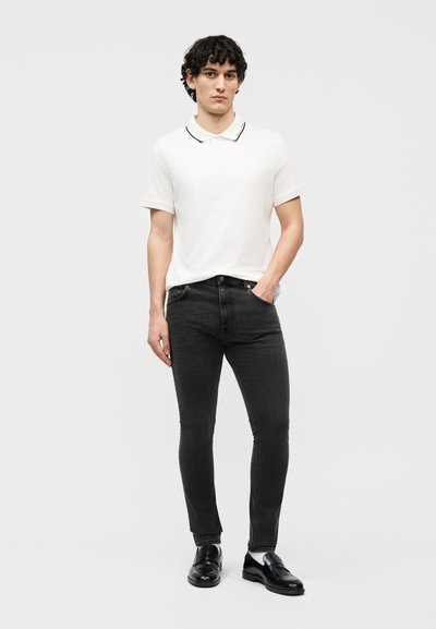 Fiatal férfi áll, fehér rövid ujjú pólót, fekete farmert és fekete bőrcipőt visel, egyszerű világos háttér előtt.