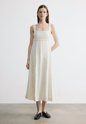 Egyenes barna hajú nő ujjatlan, fehér, midi hosszúságú ruhát visel, négyzet alakú nyakkivágással és fekete bebújós cipővel, egyszerű padlón áll.