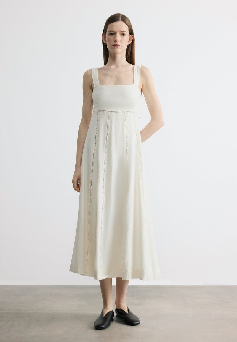 Γυναίκα με ίσια καστανά μαλλιά φοράει άσπλο μίντι φόρεμα χωρίς μανίκια με τετράγωνη λαιμόκοψη και μαύρα παπούτσια slip-on, στέκεται σε απλό δάπεδο.