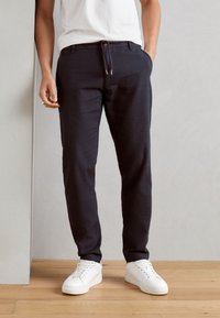 Navy tailored pants with a drawstring waist, front pockets, and a slim fit, paired with white sneakers on a wooden floor.