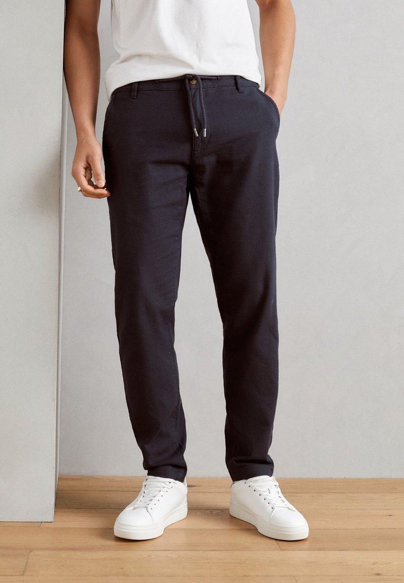 Navy tailored pants with a drawstring waist, front pockets, and a slim fit, paired with white sneakers on a wooden floor.