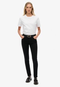 Camiseta de algodón blanca combinada con jeans ajustados negros y un cinturón negro con una doble hebilla dorada. Zapatos negros con detalles de borlas.