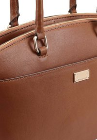 Bruine leren handtas met dubbele handvatten aan de bovenkant, zilveren hardware en een ritssluiting. Het oppervlak heeft een gestructureerde afwerking en een merkplaatje.