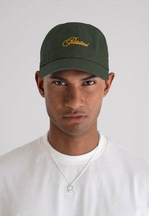 Man wearing dark green cap with yellow script, white shirt, and silver necklace with round pendant, facing forward against plain background.
