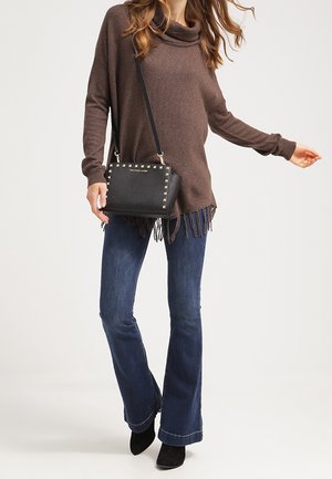Maglione marrone con scollo a cowl e dettagli in frange, abbinato a jeans a zampa d'elefante blu scuro. L'outfit include una piccola borsa nera con dettagli in borchie.