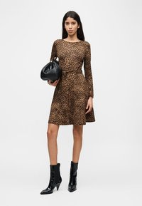 Γυναίκα με φόρεμα λεοπάρ μέχρι το γόνατο και μακριά μανίκια, κρατώντας μαύρη τσάντα και φορόντας μαύρα μποτάκια με τακούνι, μπροστά σε απλό φόντο.