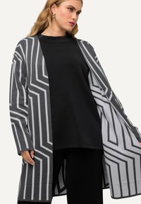 Grau und schwarzer Cardigan mit Zickzackmustern, offener Vorderseite, langen Ärmeln und weicher Textur; getragen über einem schwarzen Top und dunklen Hosen.