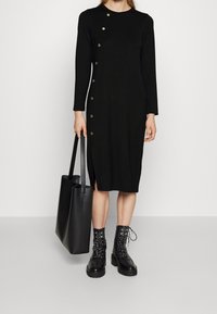 Černé dlouhé šaty s dlouhým rukávem a zlatými knoflíky po levé straně, s rozparkem na boku. Doplněné černými šněrovacími botami a velkou černou taškou.