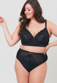 Musta pitsinen bralette, jossa on säädettävät hihnat ja pieni etuneppari, yhdistettynä korkeavyötäröisiin mustiin läpinäkyviin pikkuhousuihin. Yksityiskohtainen kukkakuvio.